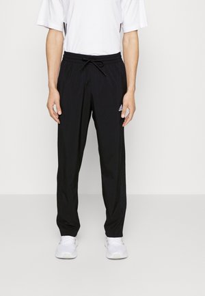 Personne portant un pantalon de survêtement noir Adidas avec une taille élastique et des baskets blanches, debout devant un fond clair et uni.