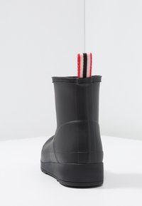 Botte en caoutchouc noir avec une surface texturée et une petite poignée à rayures rouges et blanches à l'arrière. Semelle plate, hauteur de cheville.