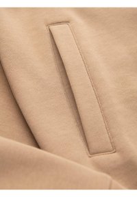 Tessuto beige con una texture liscia, caratterizzato da un elegante dettaglio della tasca e cuciture pulite, disposto in pieghe stratificate per una varietà di superficie.