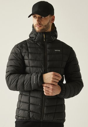 Hombre con chaqueta acolchada negra con capucha y una gorra negra, ajustándose la manga, mirando hacia un lado.