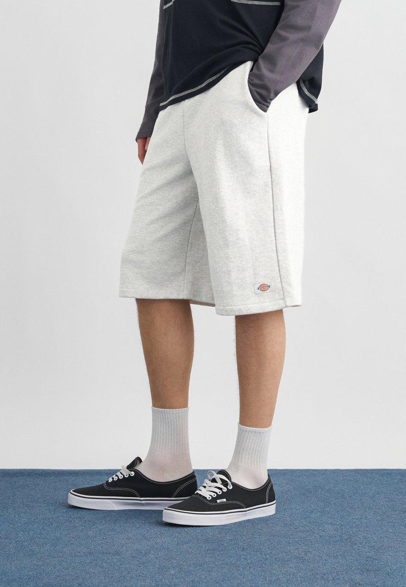 Personne portant un short gris clair arrivant aux genoux, des baskets noires avec des semelles blanches, des chaussettes blanches et un t-shirt à manches longues noir et gris.