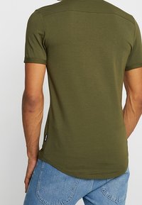 Olijfgroene katoenen t-shirt met korte mouwen, gladde textuur en een gebogen zoom. Bevat minimale merkvermelding aan de linkerkant.
