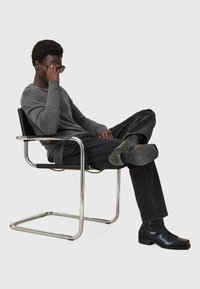 Schwarzer Metallstuhl mit verchromtem Gestell, darauf sitzt eine Person in einem grauen Langarmshirt und schwarzen Jeans, die eine dunkle Sonnenbrille trägt.