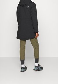 Abrigo largo negro con capucha, con un logotipo en la parte superior de la espalda. Combinado con pantalones verde oliva y zapatillas negras con detalles en menta.