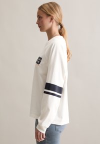 Langärmliges weißes Baumwollhemd mit einem horizontalen Marineblauen Streifen an jedem Ärmel und gesticktem Logodetail auf der Brust.