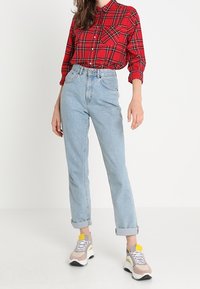 Kvinde iført en rød ternet skjorte, der er stoppet ned i højhalsede lyseblå jeans med opfoldede manchetter og beige sneakers med gule detaljer.