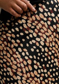 Svart tyg med stora bruna och beige abstrakta fläckar; slät textur; hand som vilar på tyget och framhäver mönsterdetaljer.