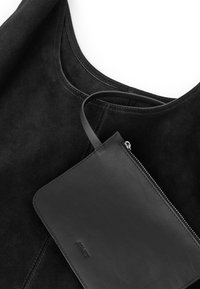 Sac fourre-tout en daim noir avec des coutures visibles et une pochette lisse en cuir noir pour poignet, fixée par une lanière avec fermeture éclair.