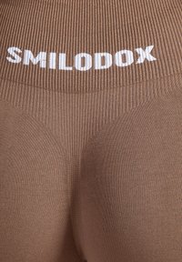 Braune Leggings mit hoher Taille und gerippter Textur sowie weißem "SMILODOX"-Logo am Bund, gefertigt aus glattem und dehnbarem Material.
