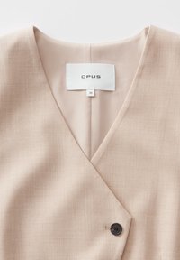 Blazer beige col V avec fermeture par un seul bouton noir et étiquette blanche indiquant OPUS, taille 36, cousue à l’intérieur du col.