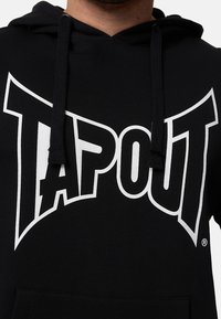 Sudadera negra con capucha que presenta un gran logo blanco de "TAPOUT" en la parte frontal, cuenta con capucha con cordón y un bolsillo tipo canguro. Tela de algodón texturizada.
