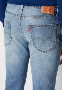Vaqueros de mezclilla azul claro con textura desgastada. Presentan dos bolsillos traseros, costuras naranjas y una etiqueta de cuero marrón con la marca.