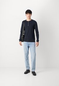 Langermet marineblå henley-skjorte med tre knapper, kombinert med lyseblå jeans og svarte slip-on sko, holder en svart crossbody-veske.