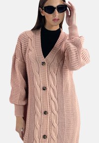 Rosa gestrickte Strickjacke mit Zopfmuster, V-Ausschnitt, Knopfverschluss und Ballonärmeln. Der Stoff hat eine gerippte Textur und große Knöpfe.