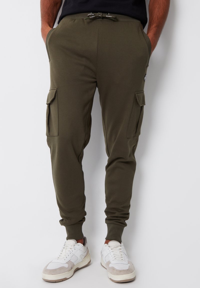 Uomo che indossa pantaloni jogger cargo verde oliva con tasche laterali e sneakers bianche, mani in tasca, in piedi davanti a uno sfondo bianco.