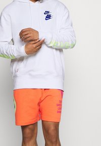 Vit hoodie med blå Nike-logotyper och grön "LOVE"-text på ärmarna, kombinerad med lysande orange shorts med stadsnamn i lila tryck.