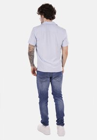 Camicia azzurra a maniche corte, design semplice, in cotone, indossata con jeans blu e sneaker bianche, tatuaggi visibili sulle braccia.