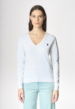 Una donna con i capelli lunghi indossa un maglione bianco a V e jeans azzurri, posando su uno sfondo chiaro. Guarda direttamente verso la fotocamera.