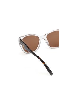 Suurikokoiset, kirkkaat aurinkolasit ruskeilla polarisoiduilla linsseillä; muovikehyksissä on läpinäkyvä etuosa ja kilpikonnakuosi temppeleissä.