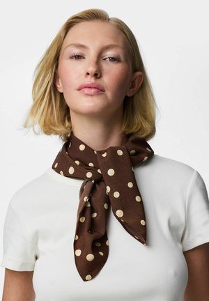 Blonde woman with short hair wearing a white ribbed top and a brown polka dot scarf tied around her neck against a white background.