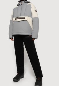 Vadderad grå jacka med en krämfärgad panel, dragkedja framtill, huva och logotypdetaljer; bärs med svarta plyschbyxor och mörka stövlar.