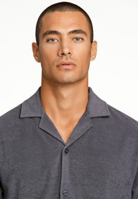 Camisa gris de botones con un tejido suave y texturizado. Presenta un cuello amplio y botones morados. Diseño simple y casual.