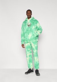 Φούτερ με κουκούλα και παντελόνι σε πράσινο tie-dye από μαλακό ύφασμα. Το φούτερ διαθέτει μπροστινή τσέπη και γραφικό σχέδιο. Το μοντέλο φοράει μαύρα παπούτσια.