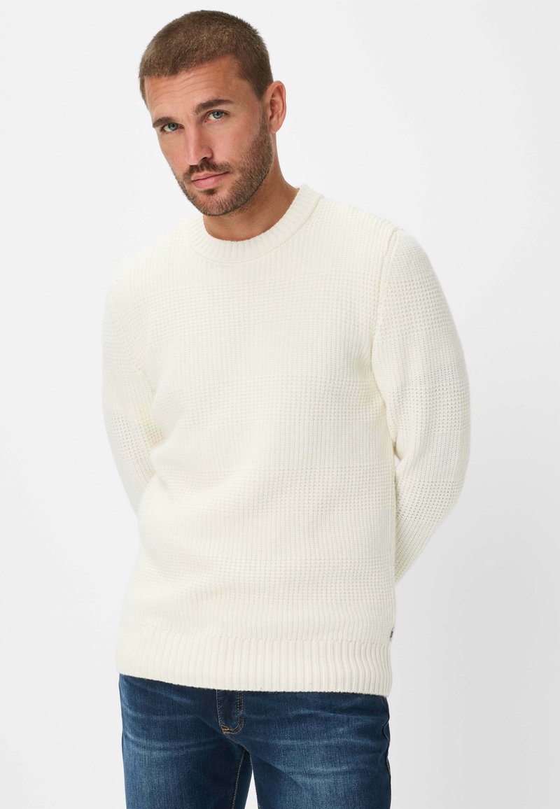 Maglione a maglia color crema con un motivo testurizzato, scollo rotondo a costine, e maniche lunghe, indossato con jeans blu scuro.