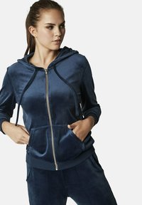 Sweat à capuche zippé bleu marine en velours lisse. Doté d'une capuche doublée, de cordons de serrage et de poches avant. Coupe décontractée avec des poignets côtelés.