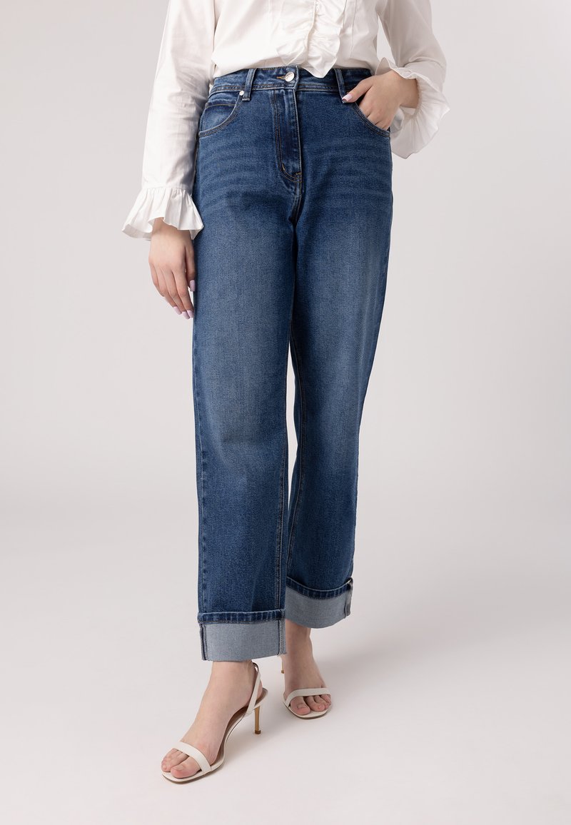 Jeans blu a vita alta realizzati in denim, con una vestibilità rilassata, orli arrotolati e una leggera scoloritura. Abbinati a scarpe col tacco color crema.