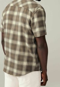 Muž v krátkém rukávu nosící hnědou a béžovou kostkovanou košili a bílé kalhoty, stojící zády k jednoduchému světlému pozadí.