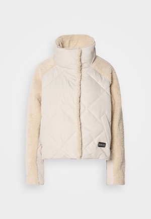Cream quilted jacket with a high collar, fluffy beige sleeves, and a visible logo patch. Soft texture, cropped design, and diagonal stitching.