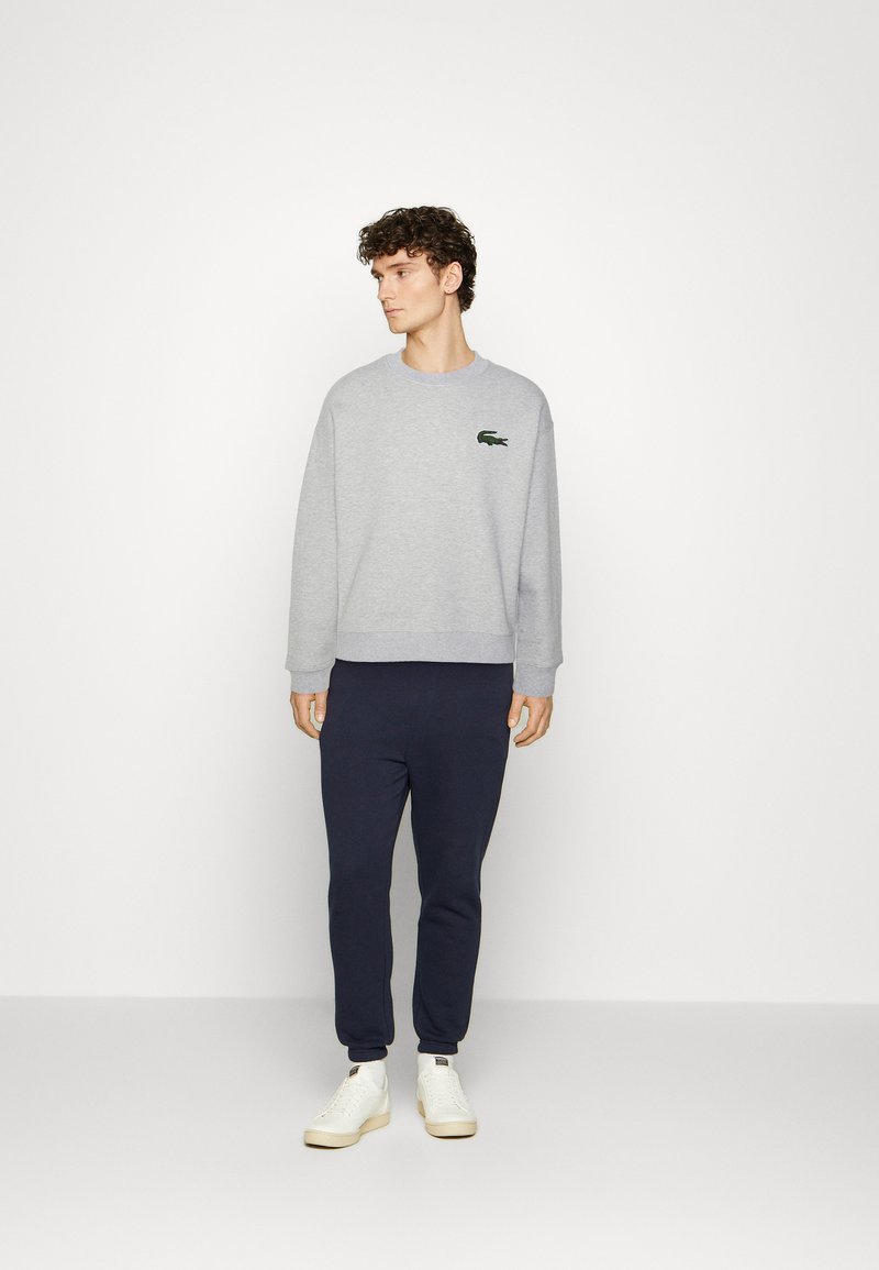 Szary bawełniany sweatshirt z okrągłym dekoltem oraz zielonym logo, zestawiony z ciemnoniebieskimi spodniami dresowymi i białymi butami; luźny krój i gładka tekstura.