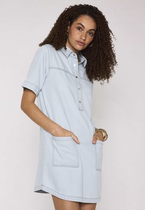 Vrouw met krullend haar, gekleed in een lichtblauwe denimjurk met korte mouwen en zakken aan de voorkant, staand met handen in de zakken tegen een effen achtergrond.