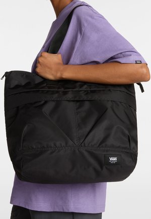 Personne en chemise violette tenant un grand sac fourre-tout Vans noir sur l'épaule, sur fond uni.