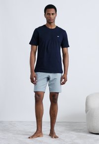 Tmavomodré tričko s krátkym rukávom a svetlé krátke nohavice so vzorom, zobrazujúce geometrický dizajn. Bosý, stojaci na mäkkom povrchu.