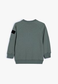 Felpa verde con una texture a coste, maniche raglan e un accento nero sul braccio sinistro. Presenta un collo a giro e polsini aderenti.