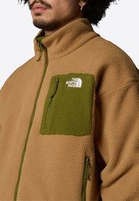 Giacca in pile color crema con tasca sul petto verde con chiusura a zip, tessuto testurizzato, colletto alto e logo The North Face in bianco.