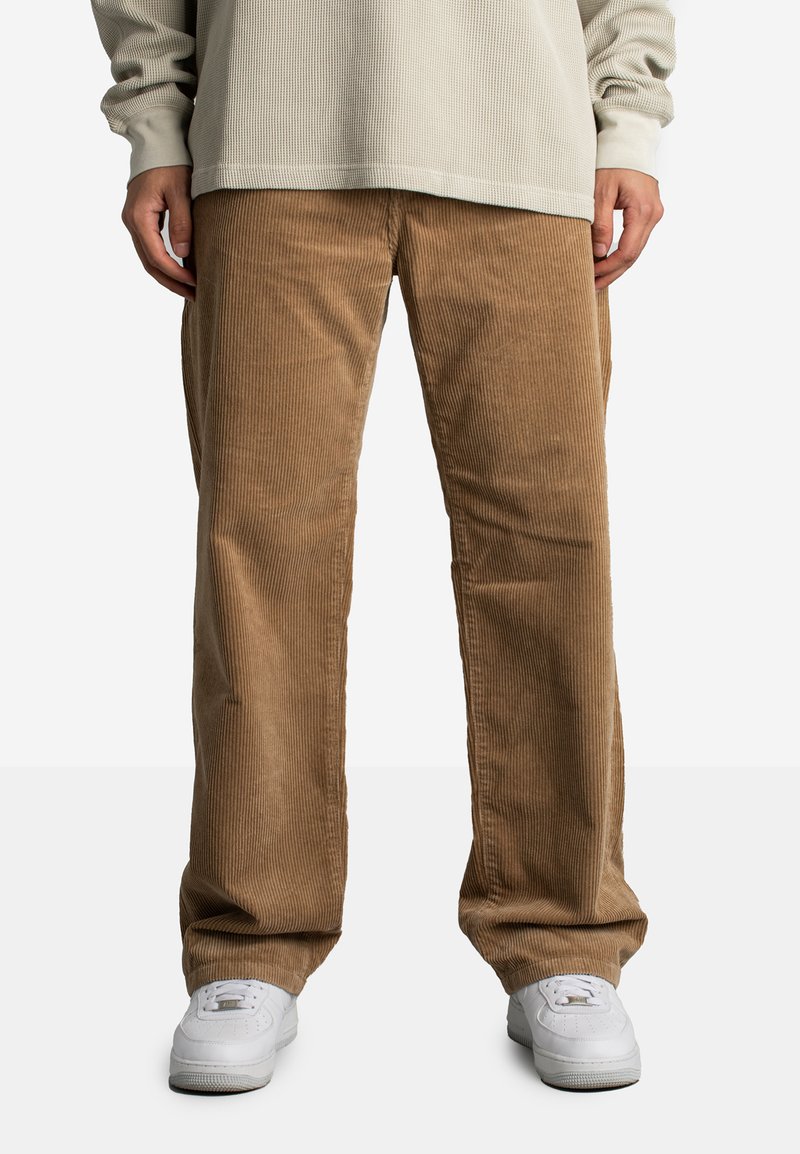 Beżowe spodnie z korduroju noszone przez osobę w jasnobeżowej, teksturowanej, długozaszywanej koszuli oraz białych trampkach, stojącą na jednolitym tle.