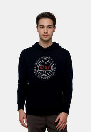 Czarna bluza z kapturem wykonana z bawełny, z okrągłym nadrukiem z napisem "GANT" w czerwonym i białym kolorze oraz "NEW HAVEN CT AMERICAN SPORTSWEAR" poniżej.