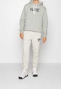 Grå huvtröja med svart "NIKE"-logotyp, kombinerad med beige joggers med svarta detaljer och en logotypmärke. Vita och svarta sneakers kompletterar outfiten.