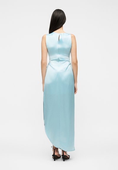 Γυναίκα με μακριά σκούρα μαλλιά που φοράει αμάνικο ανοιχτό μπλε σατινέ φόρεμα με σχίσιμο στην πλάτη και μαύρα ψηλοτάκουνα σανδάλια.