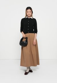 Black cardigan with buttons, tan A-line skirt, black handbag, and burgundy shoes. Model stands against a white background.