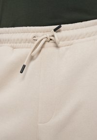 Schaumfarbene Jogginghose mit Kordelzugbund und schwarzen Akzenten, strukturiertem Stoff und Seitentaschen, die ein glattes, elegantes Design zeigen.