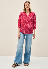 Rote Bluse mit hellrosa Blattmustern, knöpfbarem Ausschnitt, Dreiviertelärmeln. Kombiniert mit hellblauen weit geschnittenen Jeans und schwarzen Sandalen.