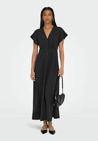 Robe maxi noire à manches courtes, col en V, devant à boutons et taille cintrée. Associée à un sac bandoulière noir et des ballerines à bout pointu.
