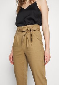 Černá saténová košilka s nastavitelnými ramínky, kombinovaná s béžovými, lehkými kalhotami s vysokým pasem, které mají svázaný pásek a záhyb v přední části.