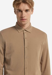 Camicia beige a maniche lunghe con chiusura a bottoni, dotata di colletto classico e tessuto strutturato. Cinque bottoni sono visibili lungo la parte anteriore.