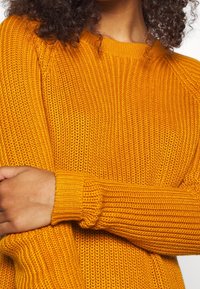 Senapsgul stickad tröja med ribbad struktur, rund halsringning och långa ärmar med åtsittande muddar.