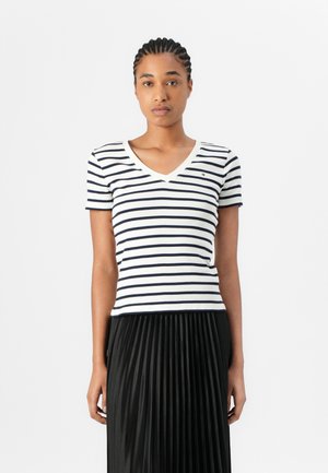 Mujer con el pelo trenzado, que lleva una camiseta blanca de manga corta con escote en V y rayas horizontales negras y una falda negra plisada.